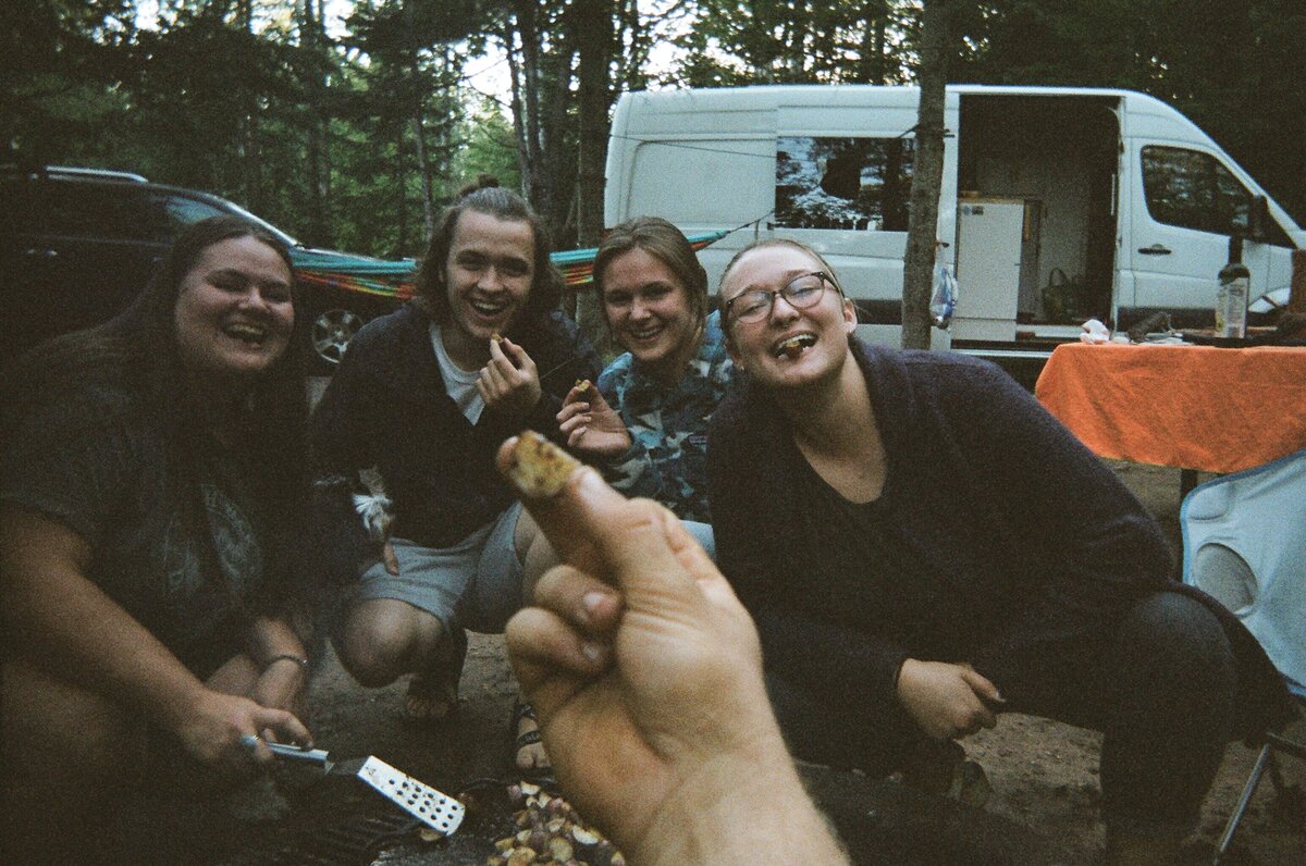 friends posing with campfire food and van behind them