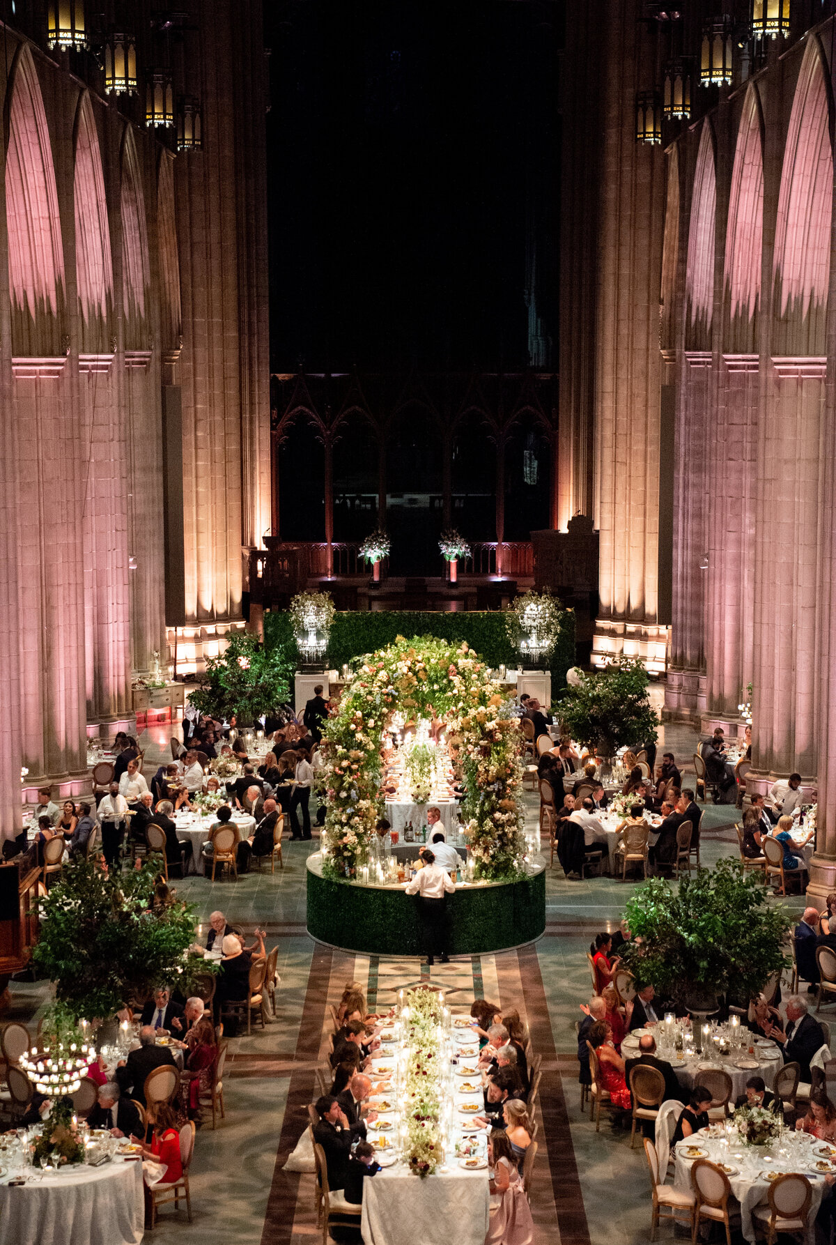 This grand wedding reception was captured beautifully at Washington National Cathedral in Washington D.C. by the talented photographer Twah Dougherty and the skilled videographers Current Films. The scene showcases an ornate, high-ceilinged hall where tables are elegantly arranged with guests seated around them. A stunning floral arch centerpiece and lush greenery add a touch of natural beauty to the space, complementing the warm lighting that highlights the intricate architectural details of the room.