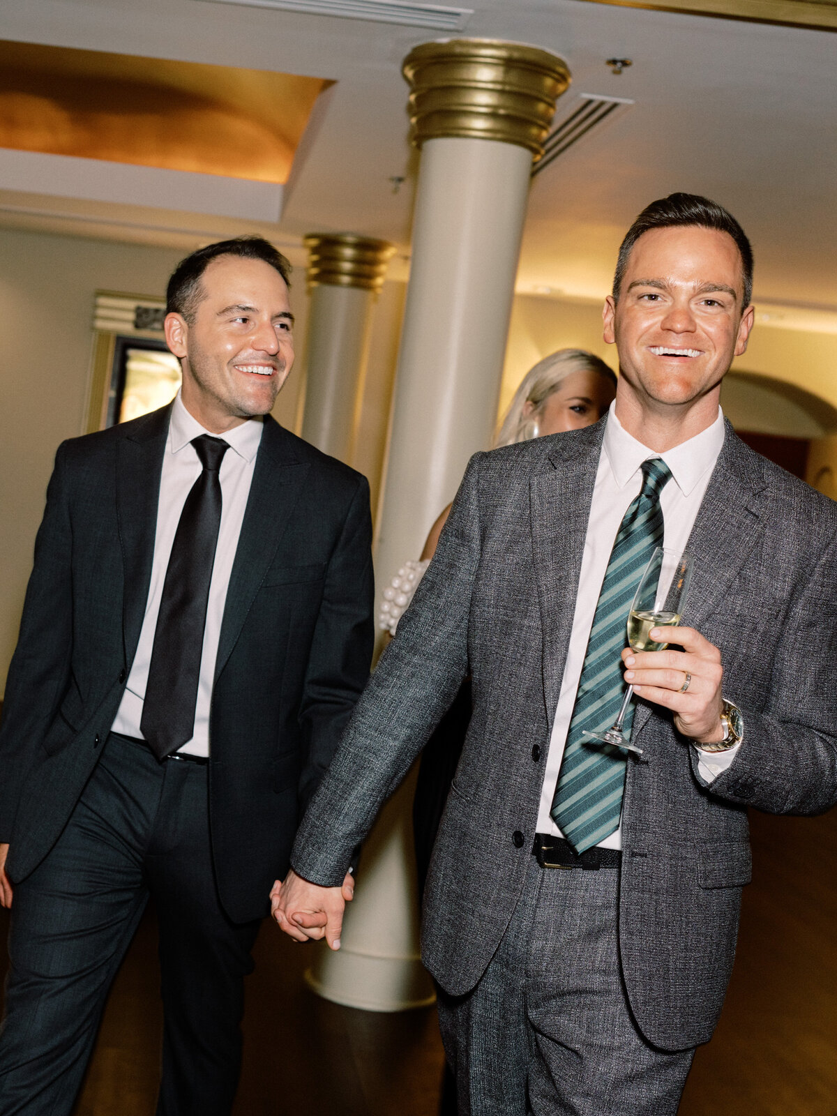 This engaging photograph depicts a stylish gay couple walking hand-in-hand through an event, showcasing their connection and the vibrant atmosphere of the gathering they are part of.