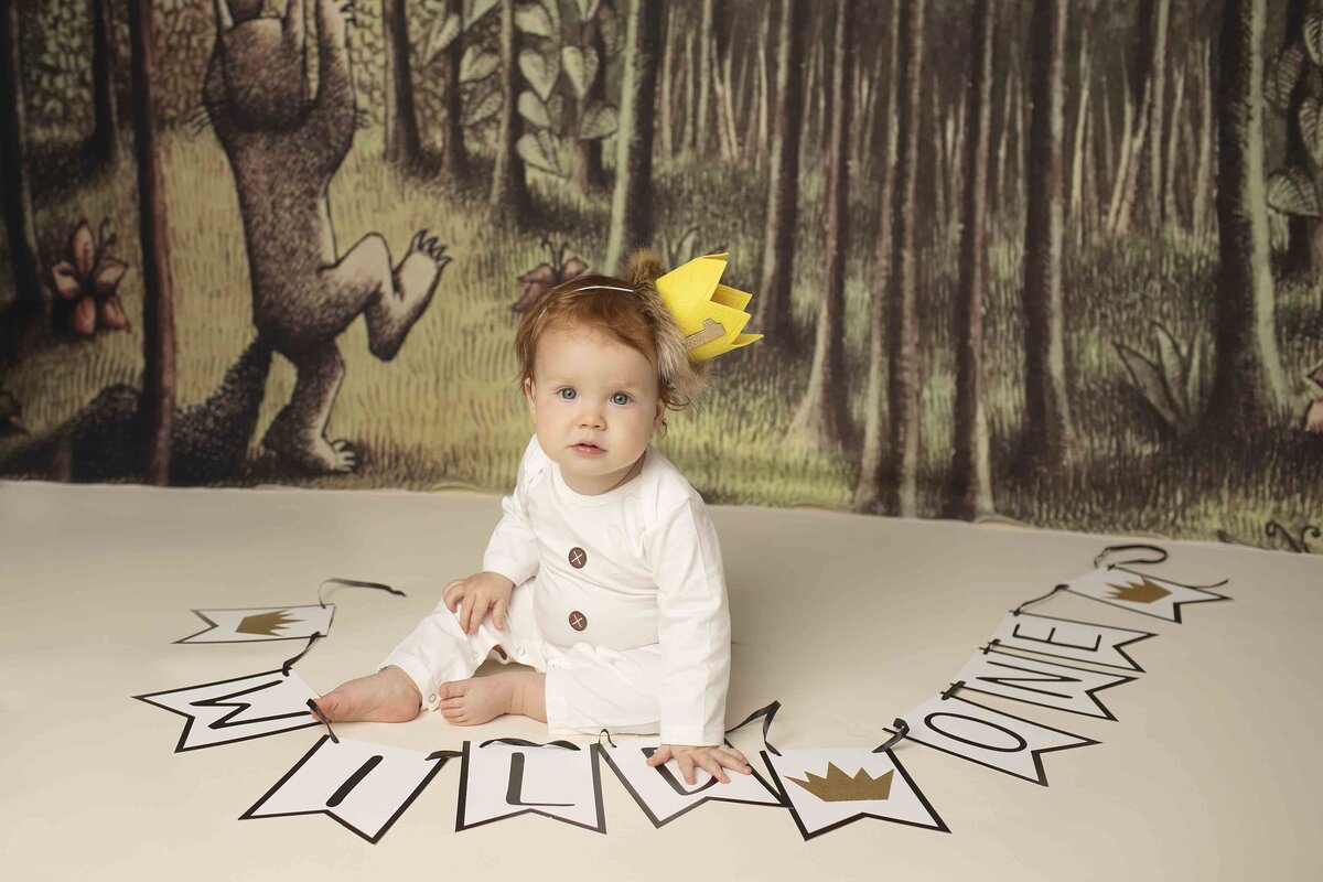 Birthday boy with red hair, wearing a Wild Thing crown, sitting next to a Where the Wild Things Are themed backdrop