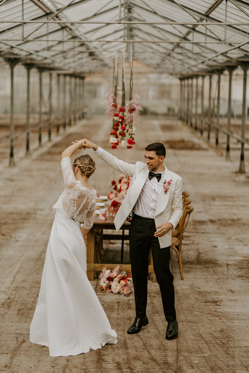 Mariée tournoyant dans une danse sous le bras de son mari sous une immense structure de métal et de verre. Table de mariage et fleurs suspendues en arrière plan. Photo capturée lors du workshop photographie de mariage.