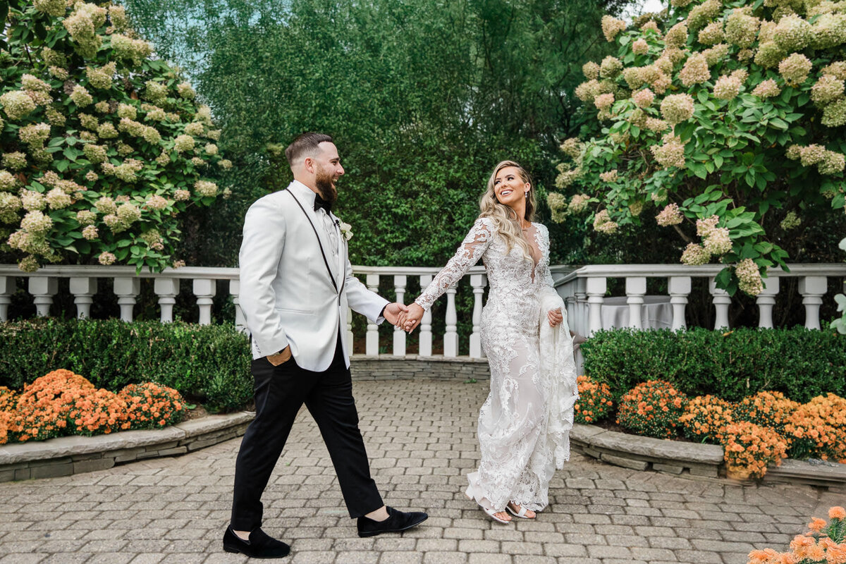 Bride and groom walk hand in hand through a garden terrace surrounded by hydrangeas, smiling at each other during a candid wedding moment.