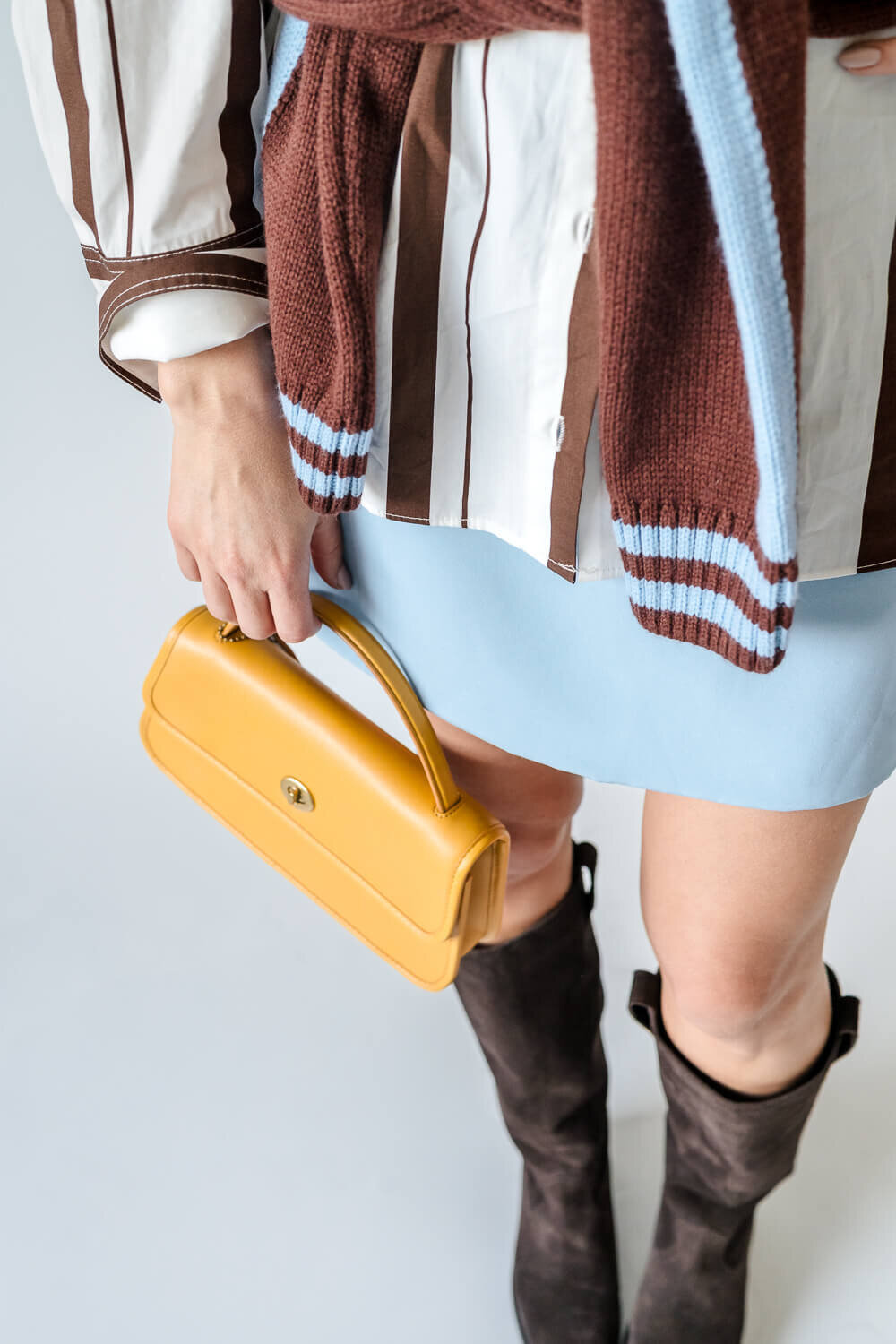 Close-up of woman holding yellow handbag, wearing brown knee-high boots and striped shirt.
