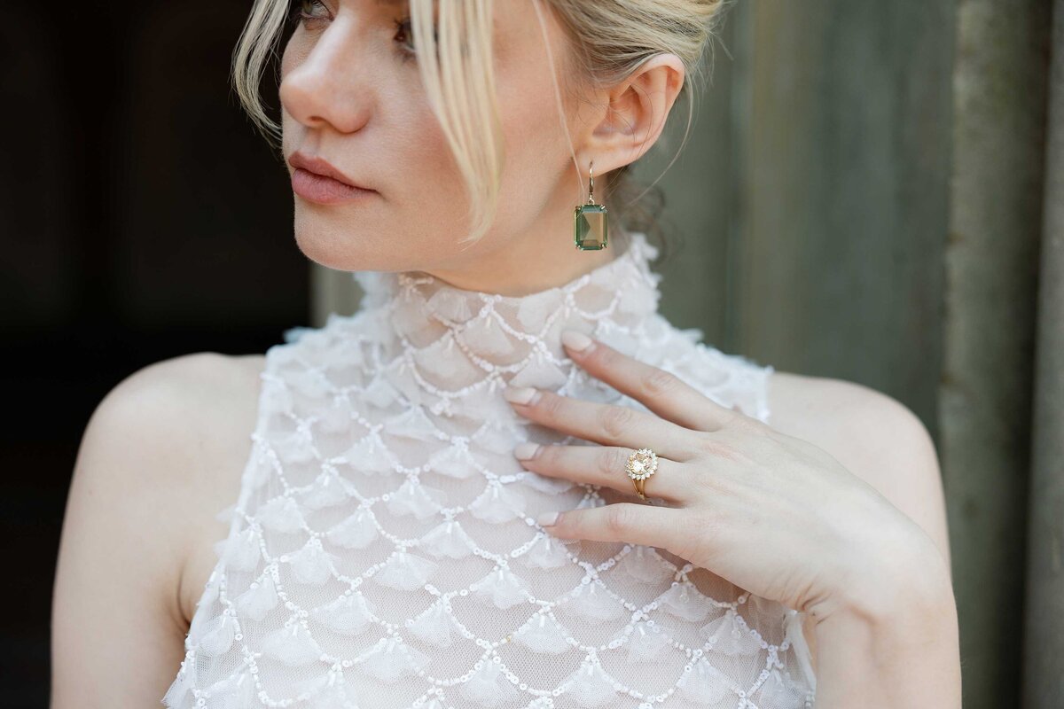 Bride looks over her shoulder while highlighting her engagement ring. She's wearing a high neck, sleeveless gown.