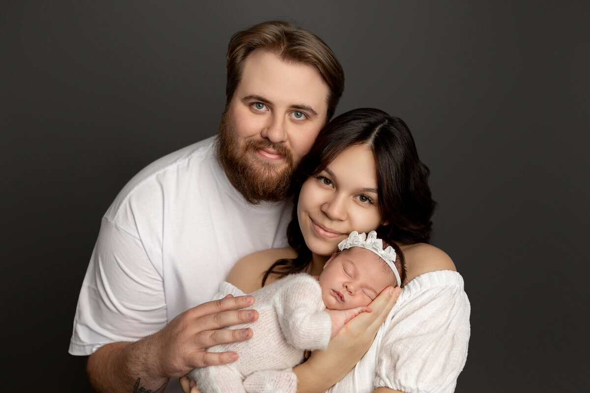 Mom and dad holding their newborn daughter in a cozy white knit outfit with a bow headband, posed together against a dark studio backdrop. Captured by Inland Empire newborn photographer Kaitlyn Dawn Photography.