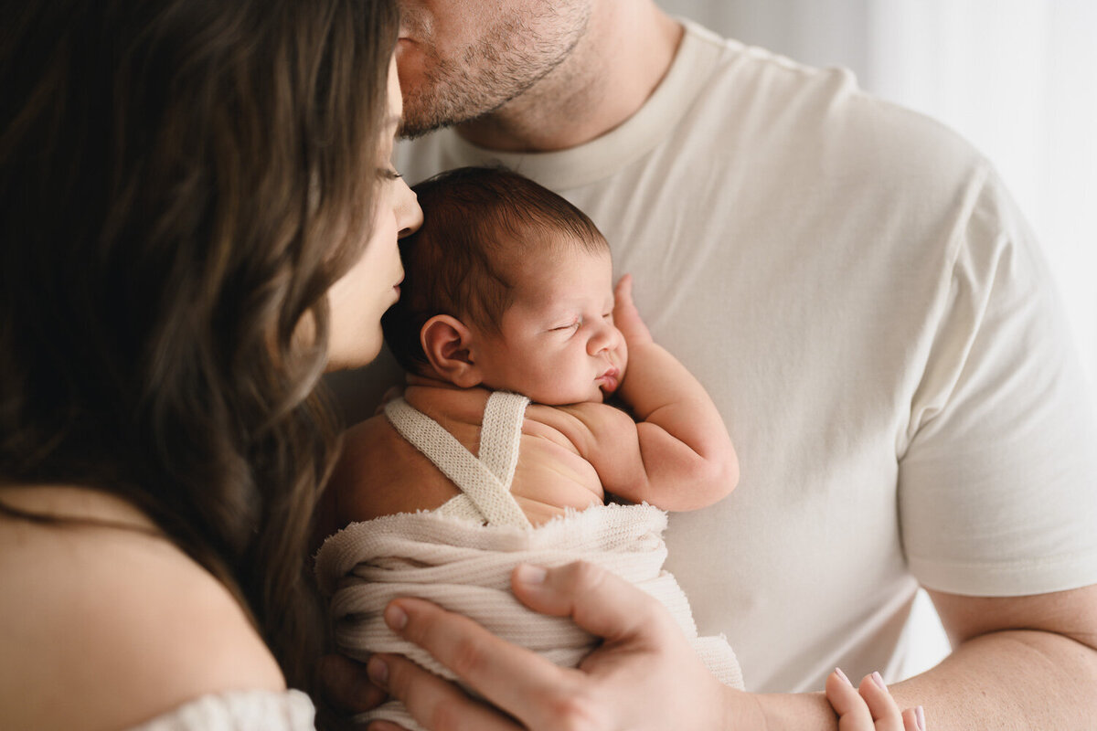 Image of father holding newborn baby on chest, with mother kissing baby, and father kissing mother in a sweet moment during their newborn photo session in Sydney