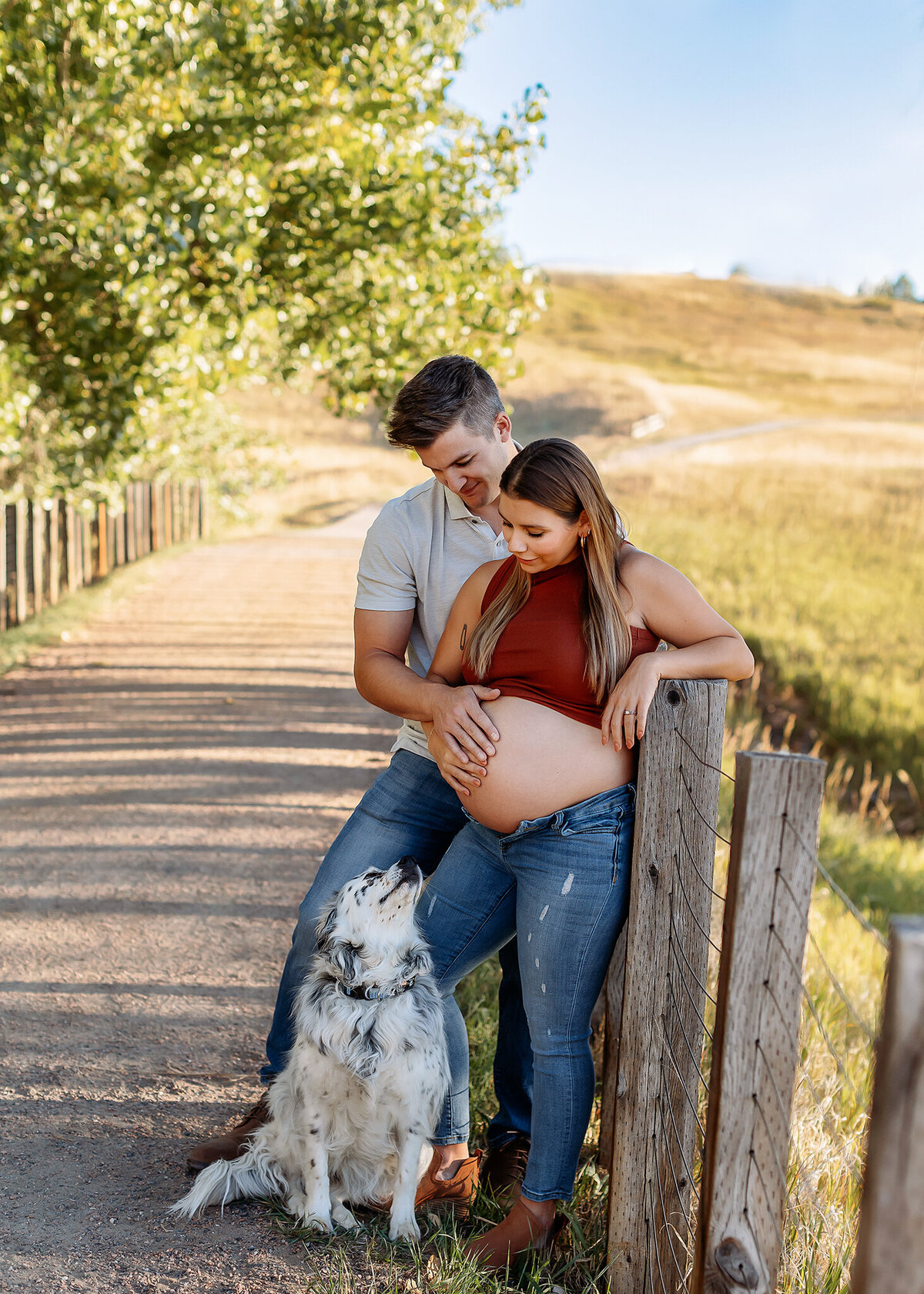 A couple stands in a golden Colorado field, eyes locked with excitement for their growing family in this romantic couples maternity photo.with their dog playing around them