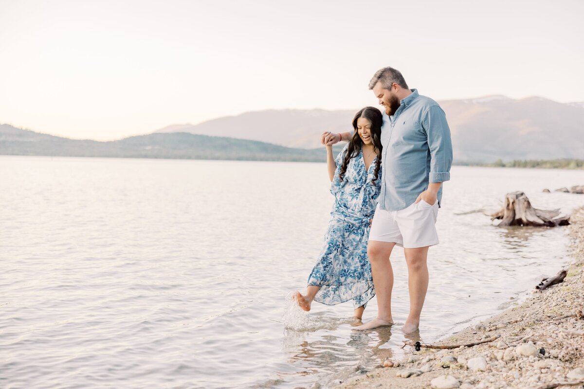 A man and woman are walking arm in arm. The woman has on a blue dress and is splashing her toes in the lake while her partner smiles at her. Taken by a local Breckenridge Photographer