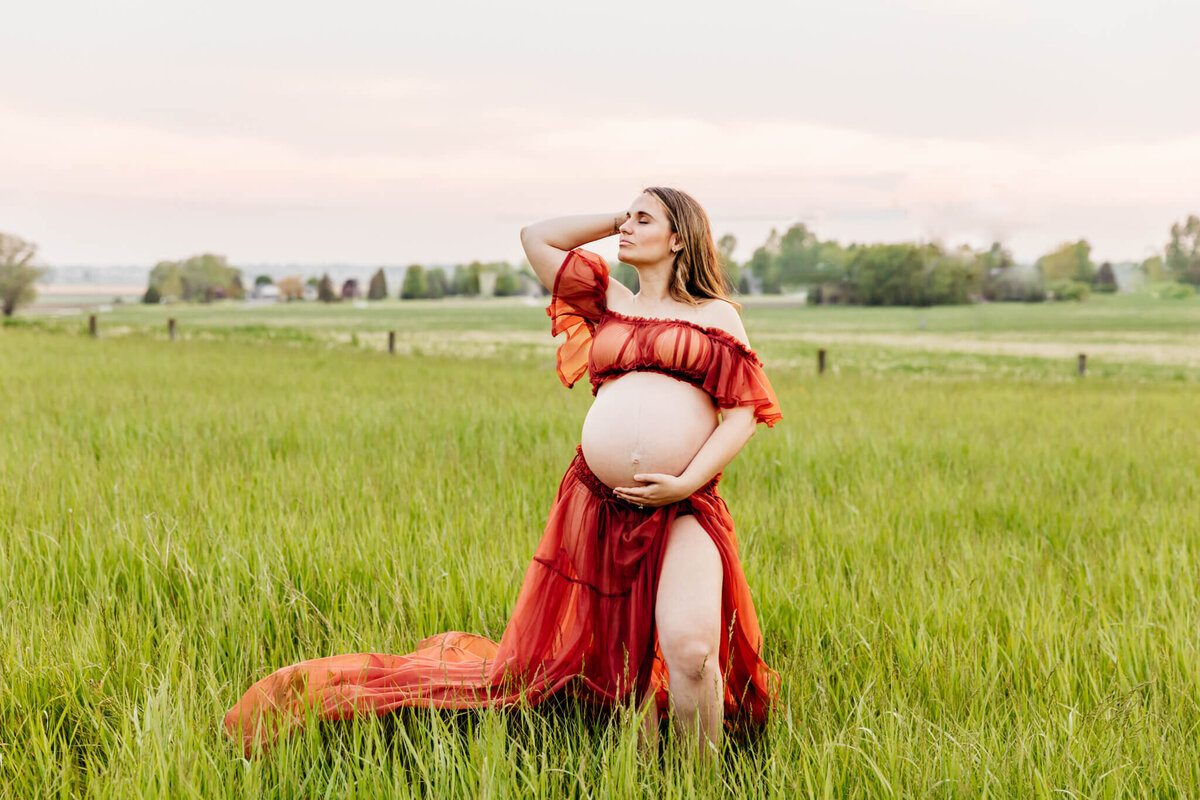 pregnant woman in an orange two piece dress closing her eyes as she holds her baby bump and puts her hand in her hair during her maternity photo session