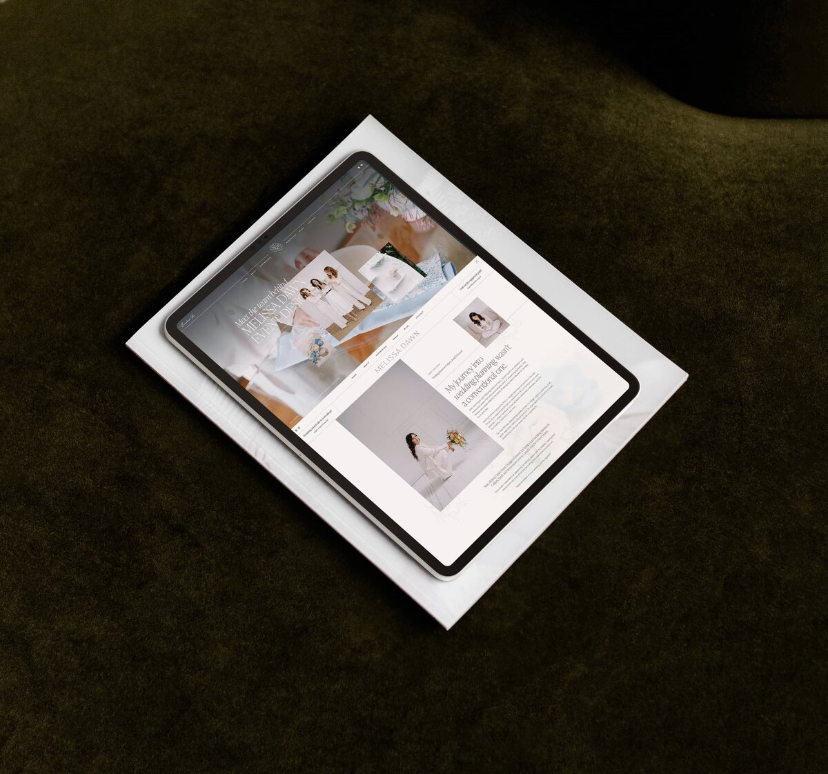 A tablet displays a stylish wedding planner website on top of a closed white book, placed on a dark green surface.