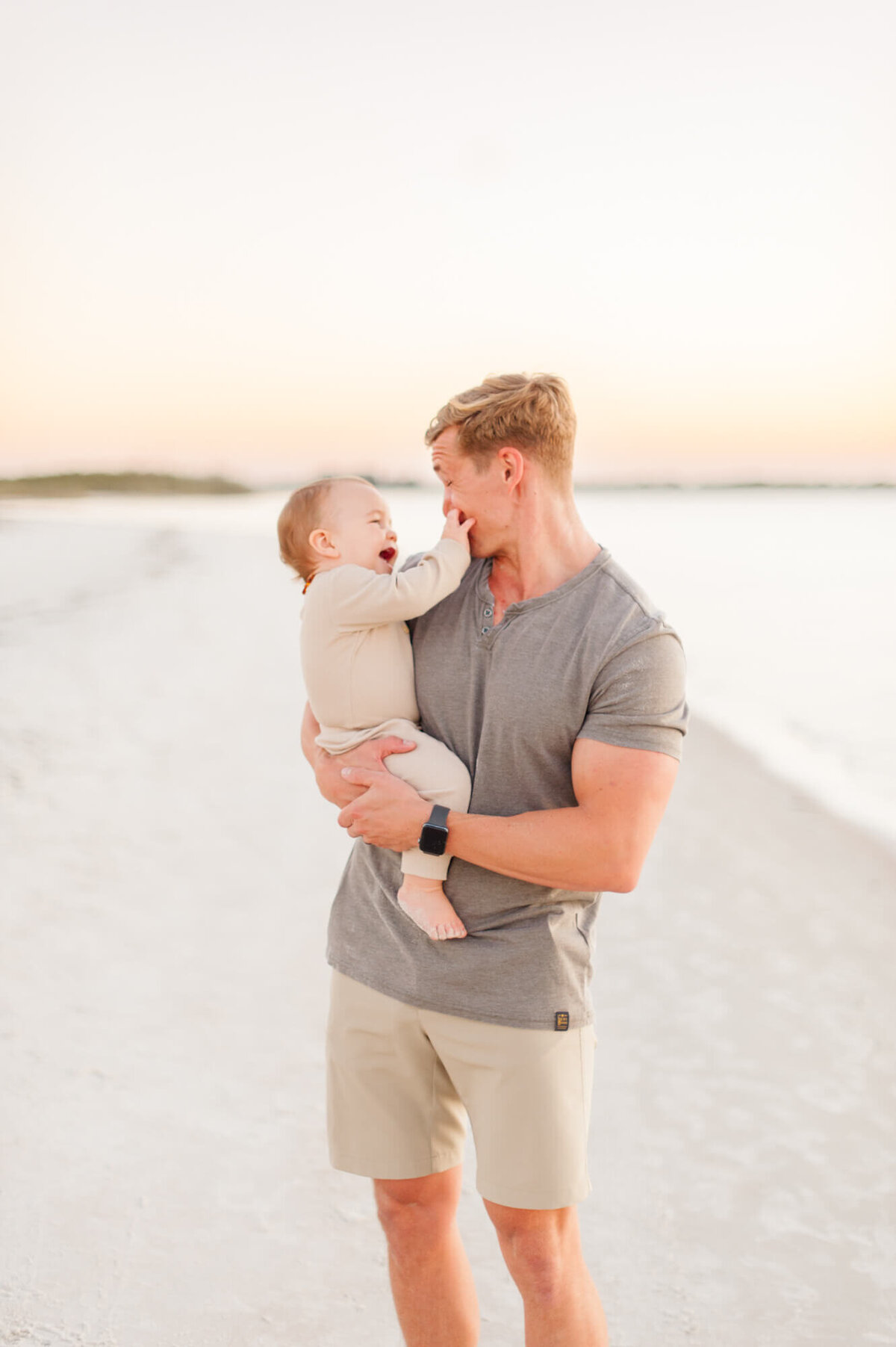 Son put his hands on dads mouth while they smile and laugh on the beach during their family portraits