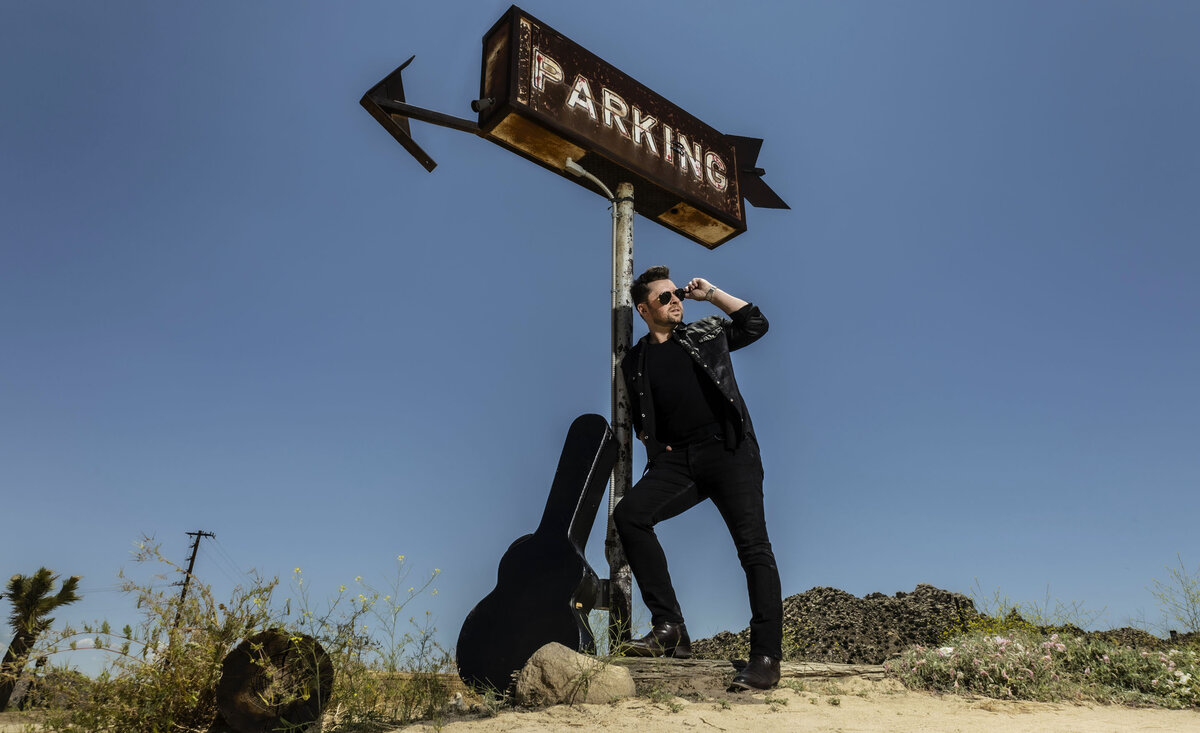 Country musician portrait Shawn Austin leaning against tall old parking sign with guitar case wearing black denim