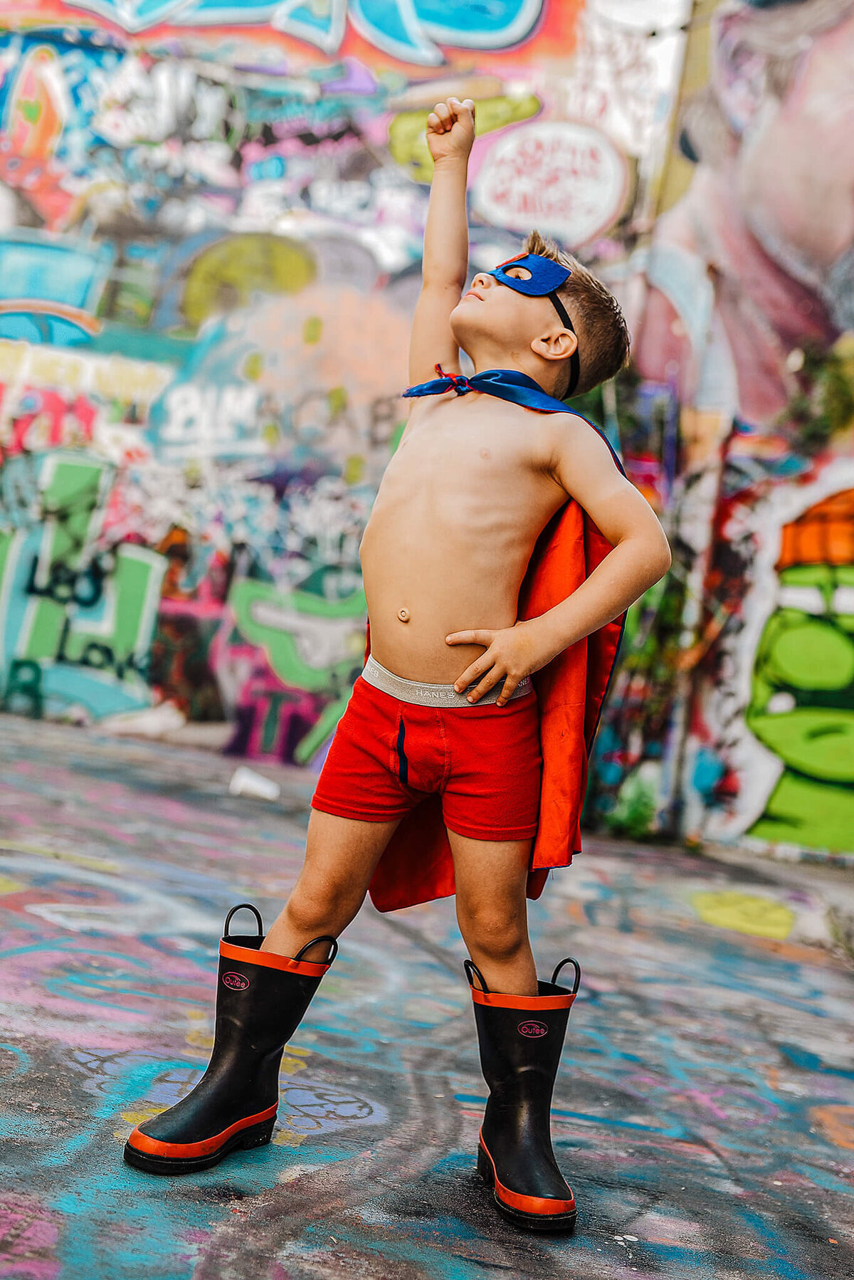 Little boy in red underwear and black boots wearing a superhero cape and mask with his fist to the sky in graffiti alley near MICA in Baltimore Maryland