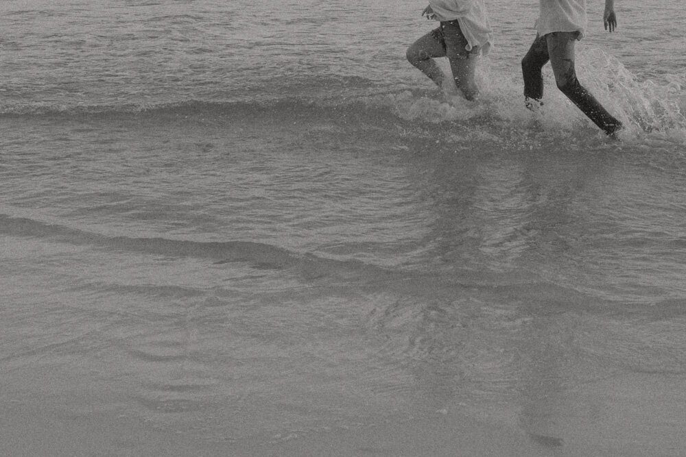 A mother and boy run through the ocean in a black and white photo with the focus on their lower body in Santa Rosa Beach, FL. 