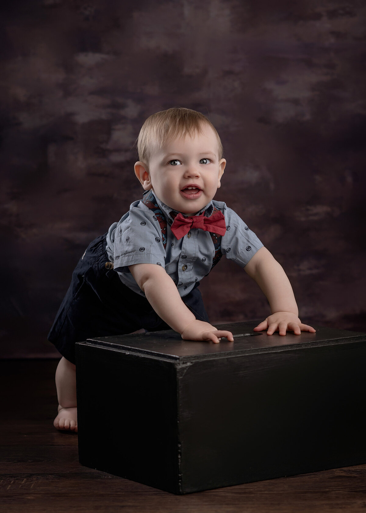 Toddler in a button up and bow tie leaning on a black box against a grey background.