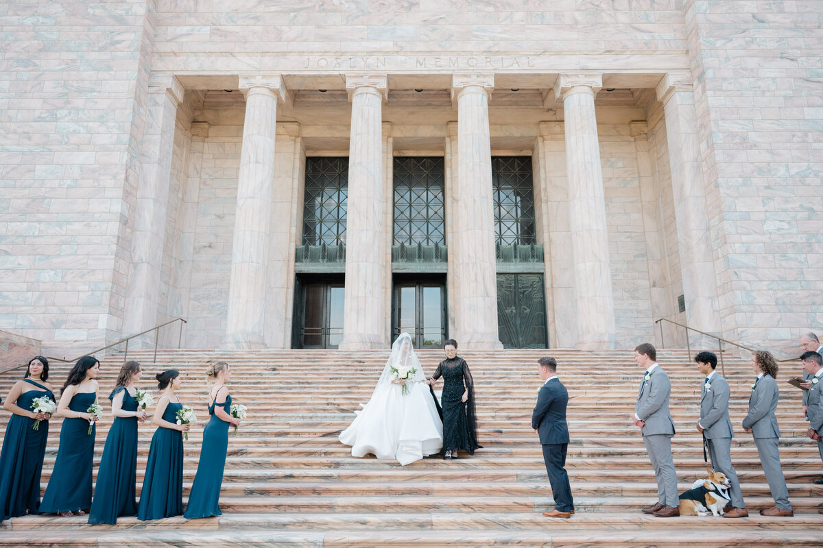 Black and white photo of a bride wearing a strapless dress with a bow and groom with a black tie tux walk hand in hand. Photo by Anna Brace, an Omaha wedding photographer.