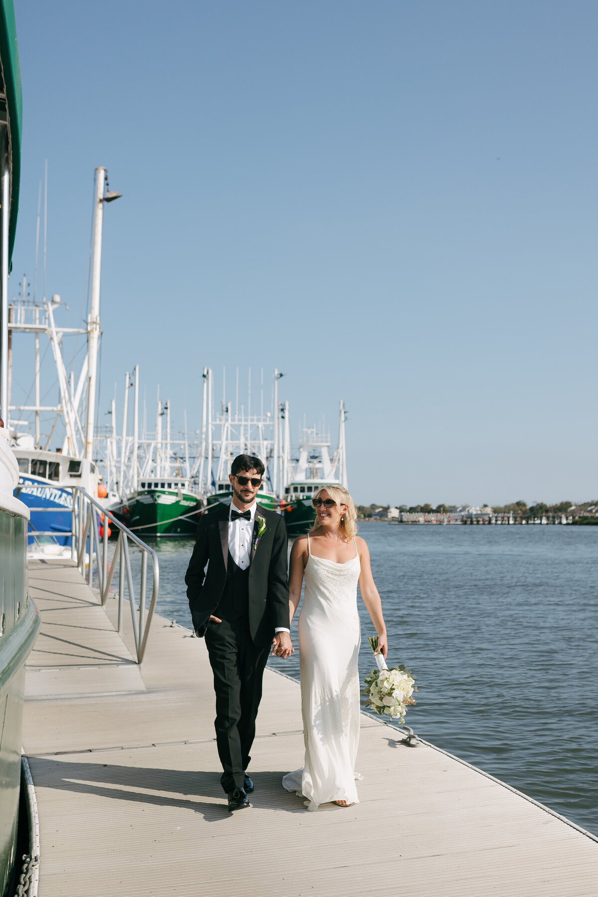 A bride and groom holding hands walking on a dock with sailboats behind them in Cape May, New Jersey.