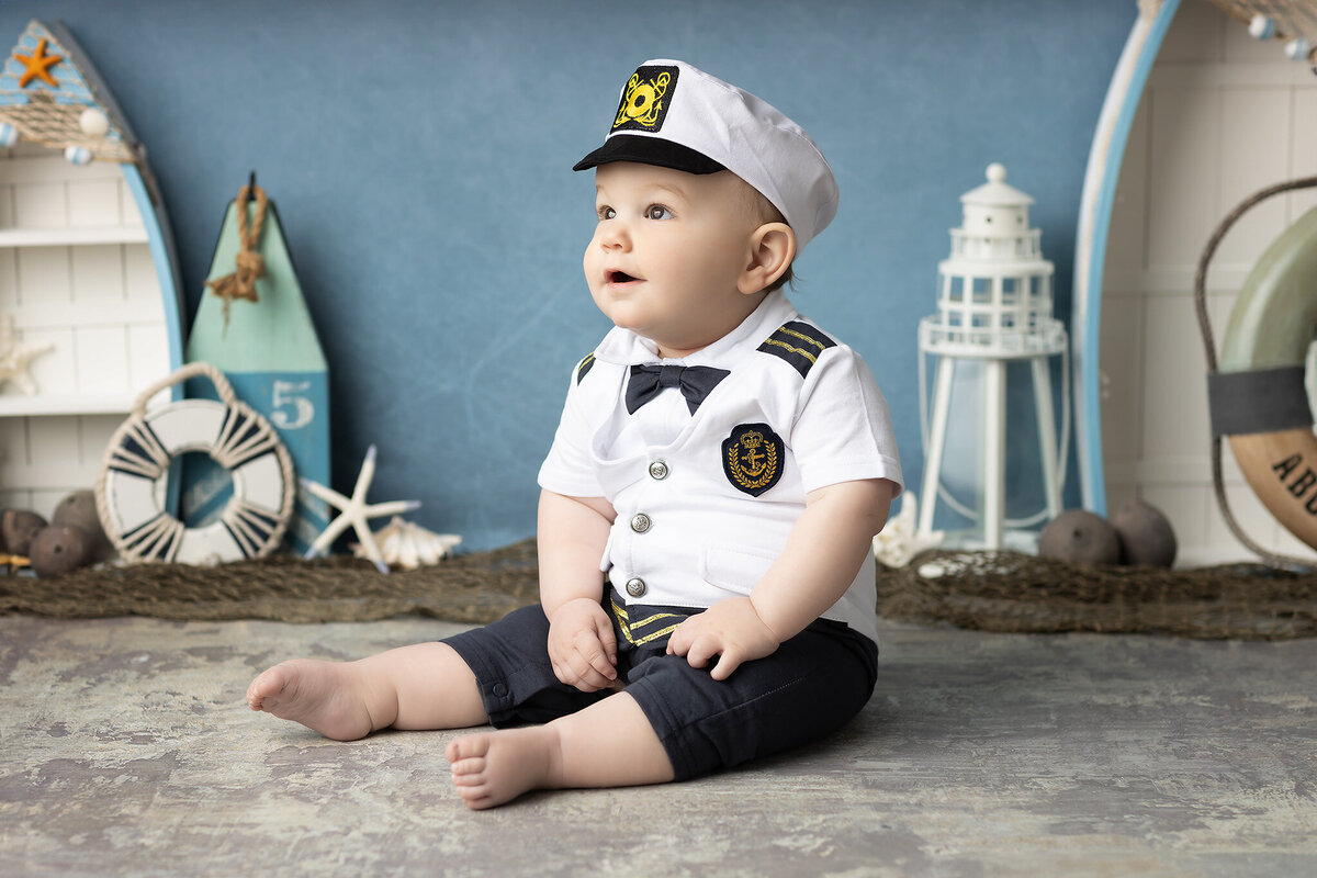 Baby boy in a white captain outfit and sailor hat during a nautical-themed photoshoot with coastal props, taken by a DFW milestone photographer.