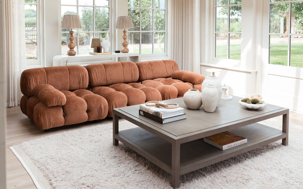 A cozy living room with large windows features a plush, rust orange tufted sofa, a light-colored rug, and a wooden coffee table adorned with decorative items and books. Two large, neutral lamps sit on a console table behind the sofa, and greenery is visible outside.