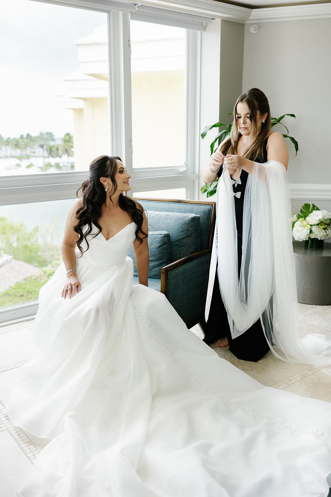 A sweet moment of the bride seated as her bridesmaid adjusts her veil, with the light streaming in from the window at Ocean Reef in Key Largo, captured by Claudia Amalia Photography during Ashley and Cameron's micro wedding.