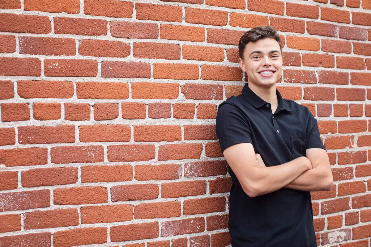 A person with short brown hair, wearing a black polo shirt, stands smiling with arms crossed against a red brick wall.