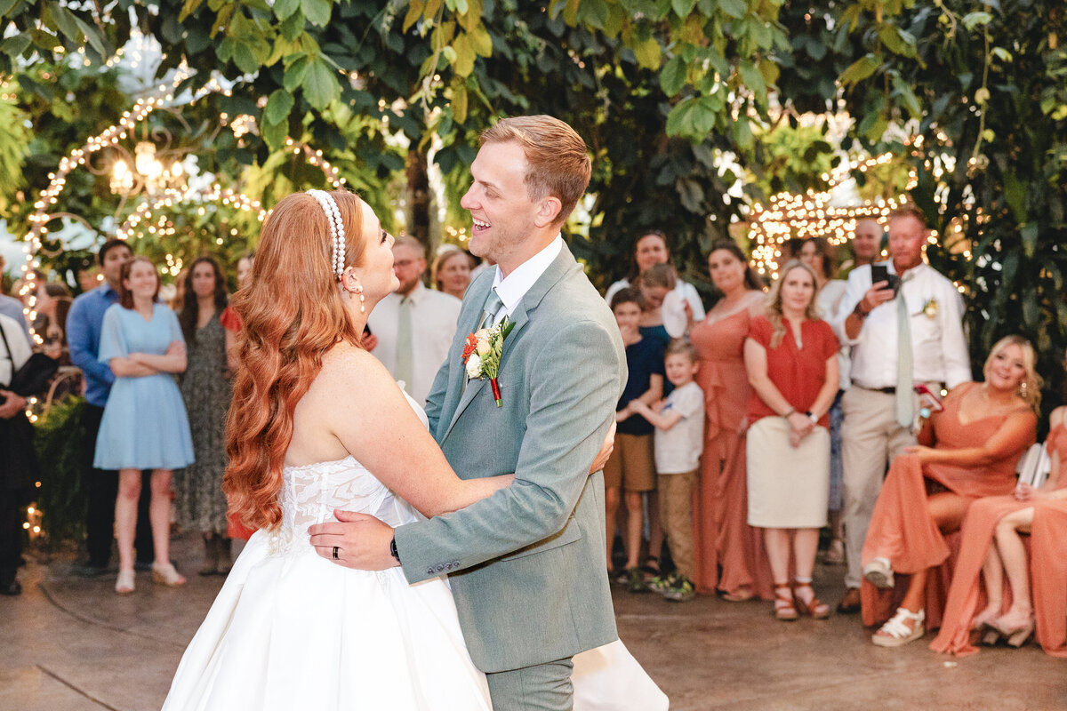 The bride and groom share their first dance under twinkling lights and lush greenery. Guests surround them, watching with smiles.
