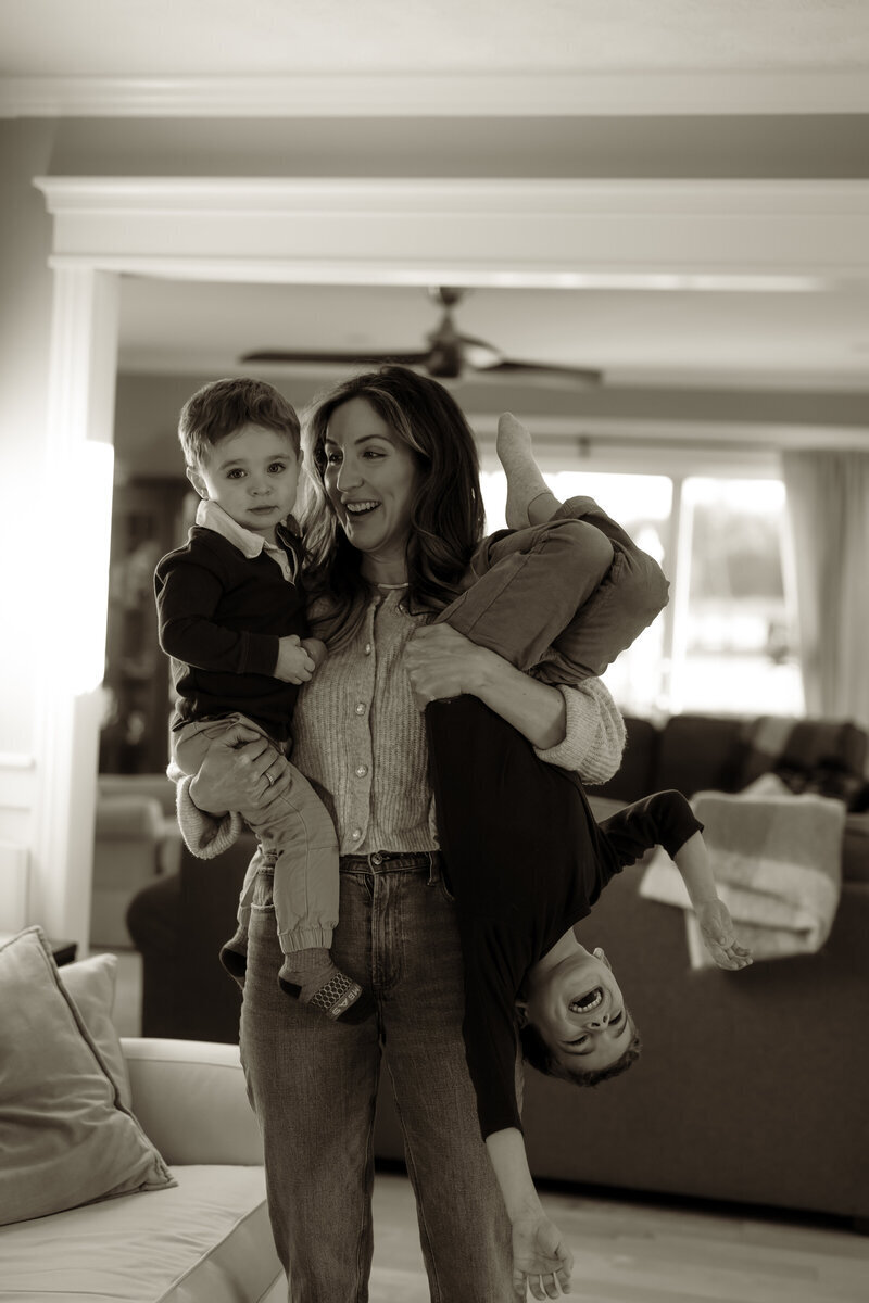 In a black-and-white scene, a joyful woman smiles warmly while cradling one child in her arms, as another dangles playfully from the other. The cozy living room setting hints at the artful touch of a New England Portrait Photographer.