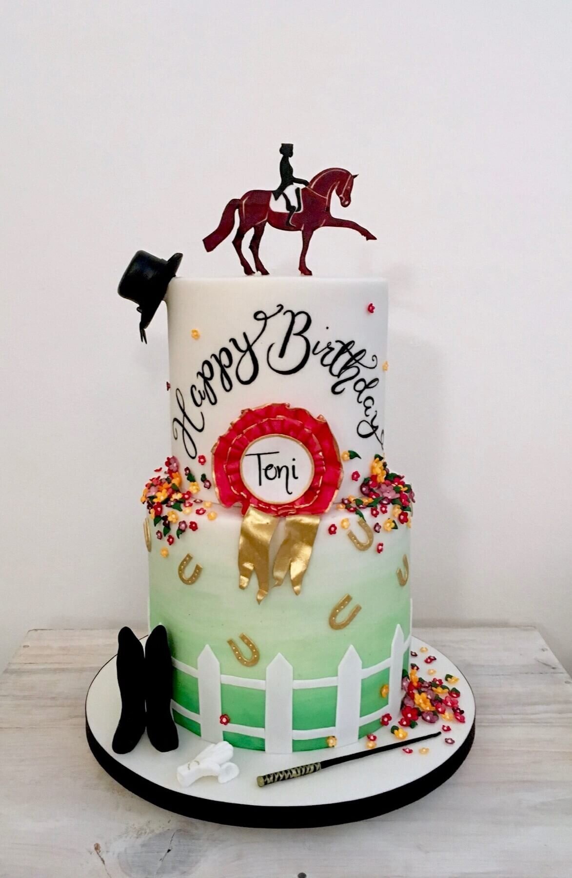 A two tier birthday cake with a horse rider theme showing boots, gloves, a hat, and award and more