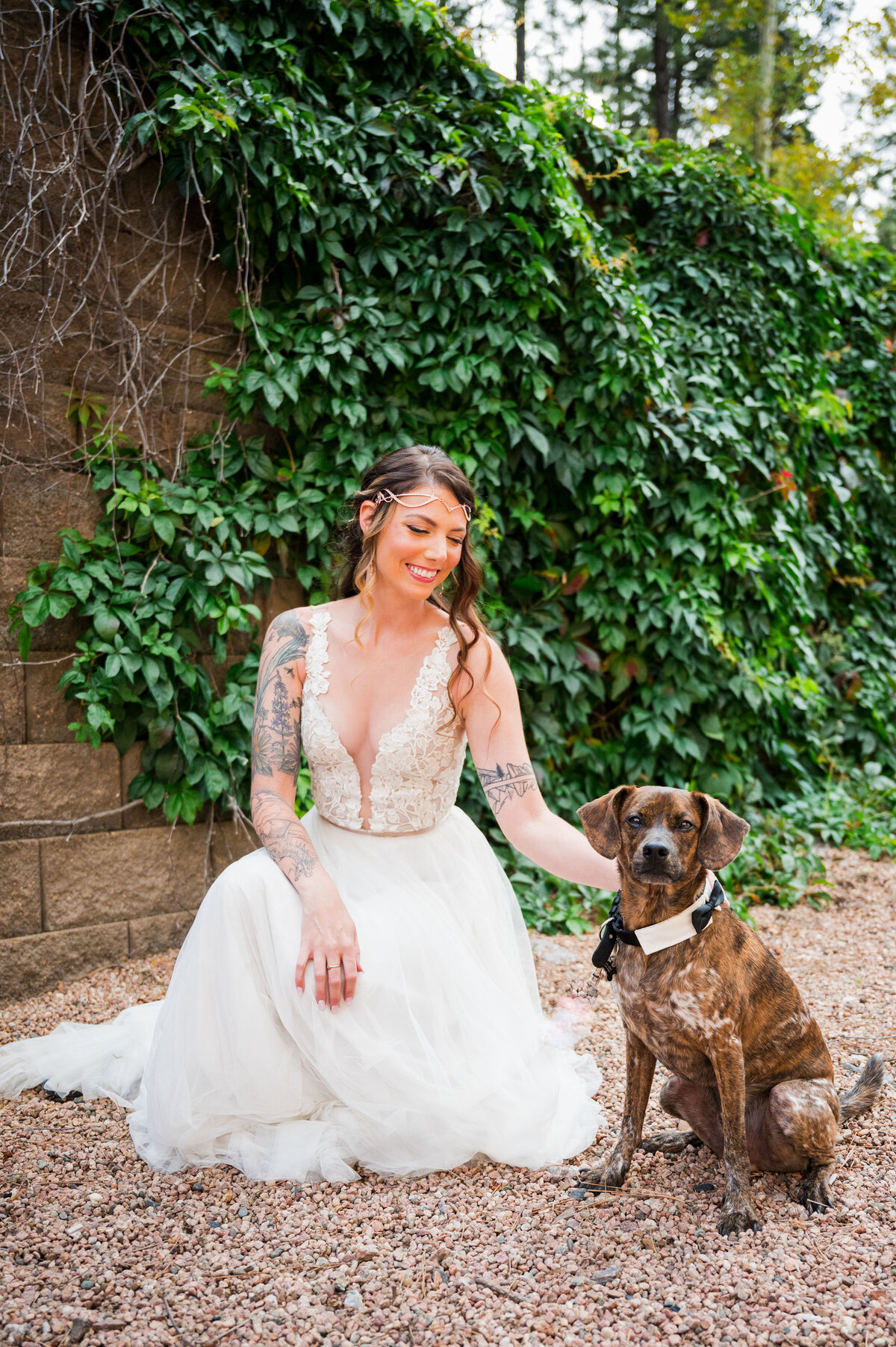 A bride kneels on the ground to pet her puppy.