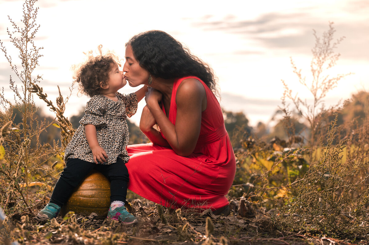 Mom kissing baby sitting on a pumpkin in a pumpkin patch