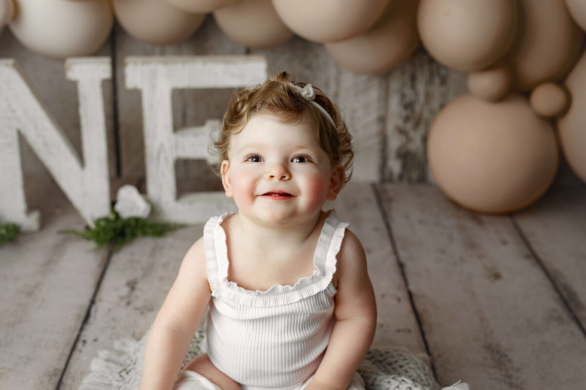 toddler smiling at the camera with a white outfit and nuetral balloons behind her on a wooden white backdrop