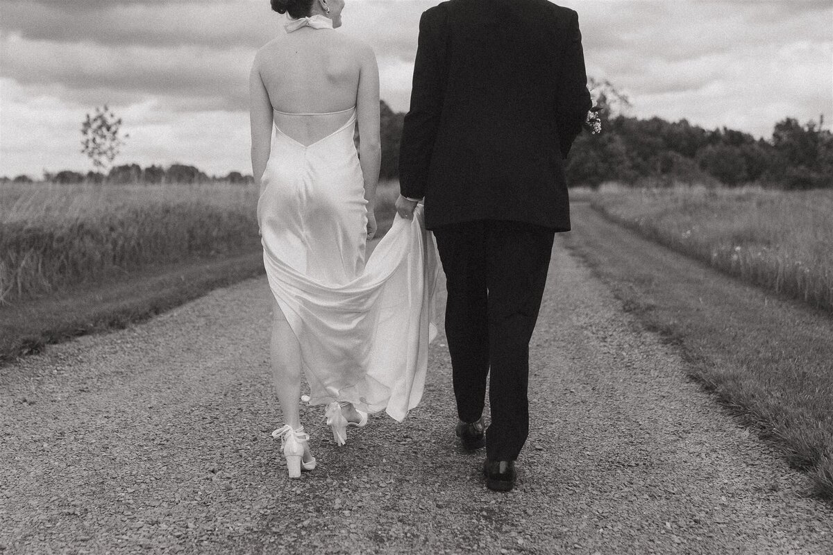 Bride holds up her dress and groom holds bridal bouquet as they run through meadow together.