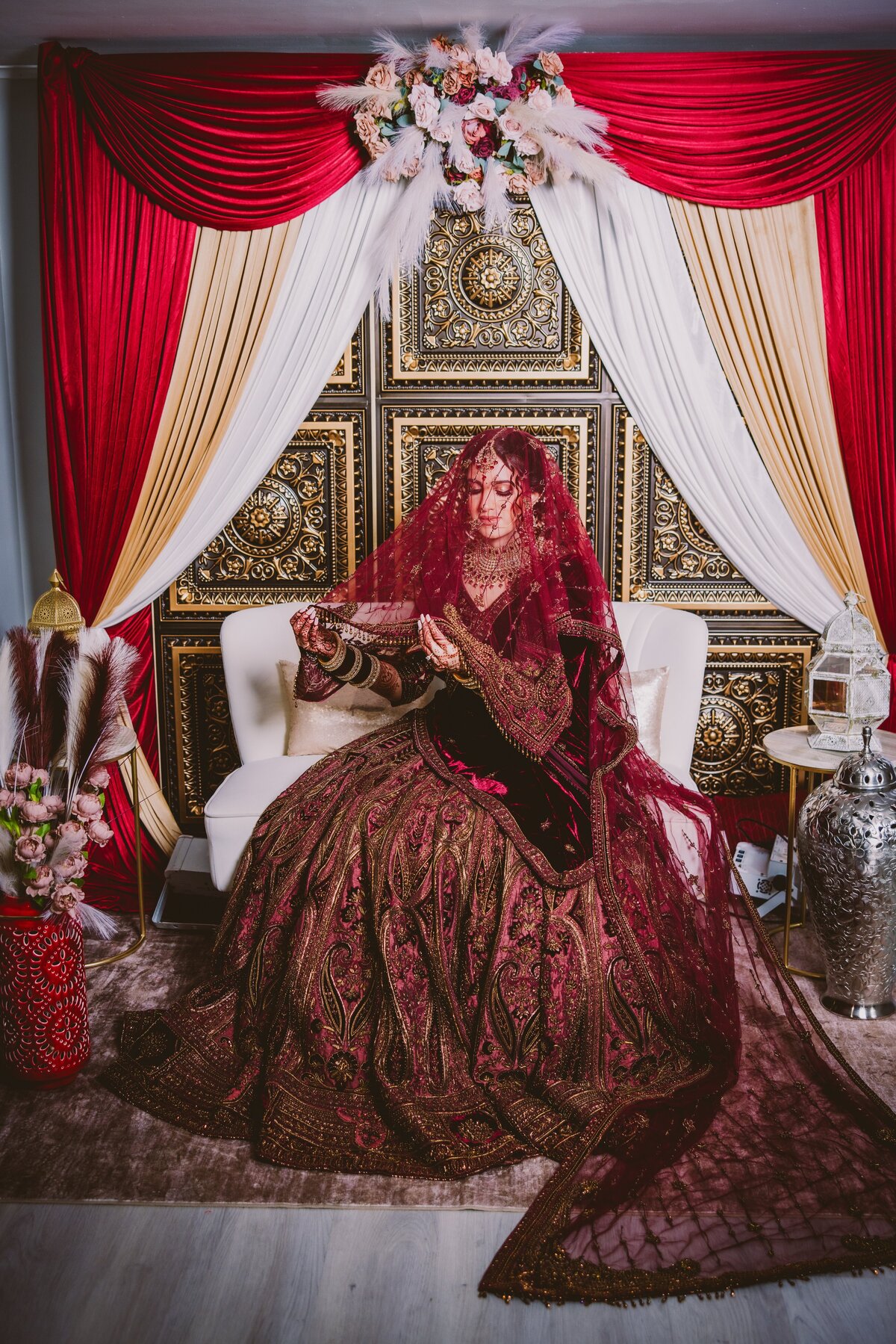 A sikh bride with a red dupatta veil over her head sits on a sofa in front of a gold backdrop
