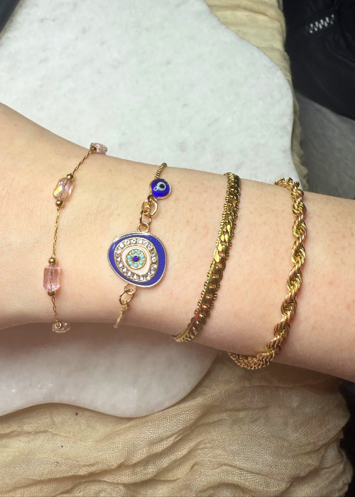A wrist adorned with a stack of five gold bracelets, including a large evil eye charm and pink crystal beads, showcasing a bold and stylish permanent jewelry look.