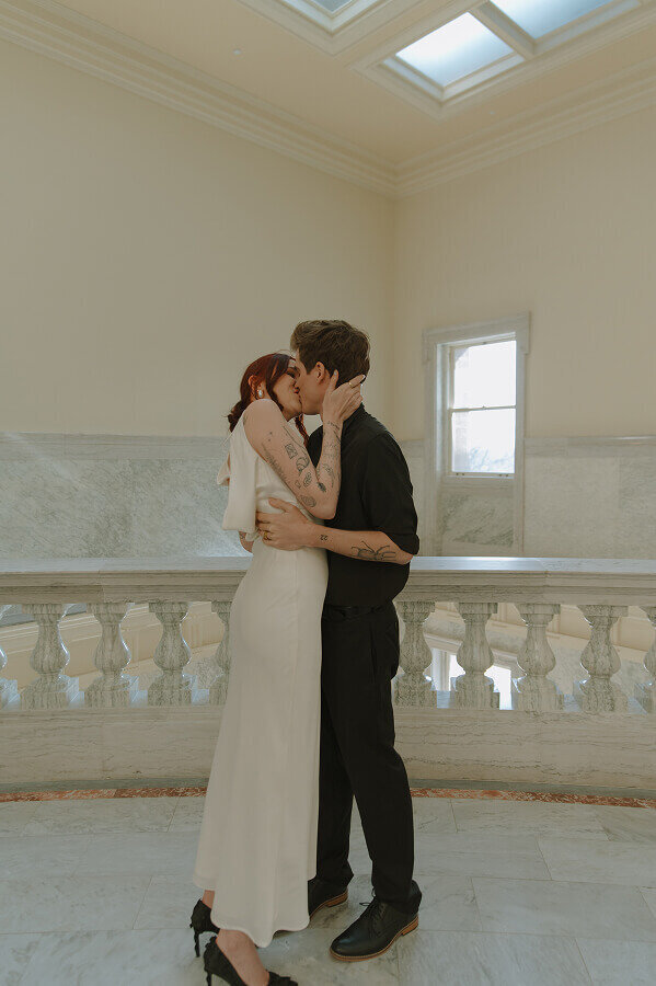 A man and woman kiss on their elopement day. They're standing inside of a building with an all white interior.