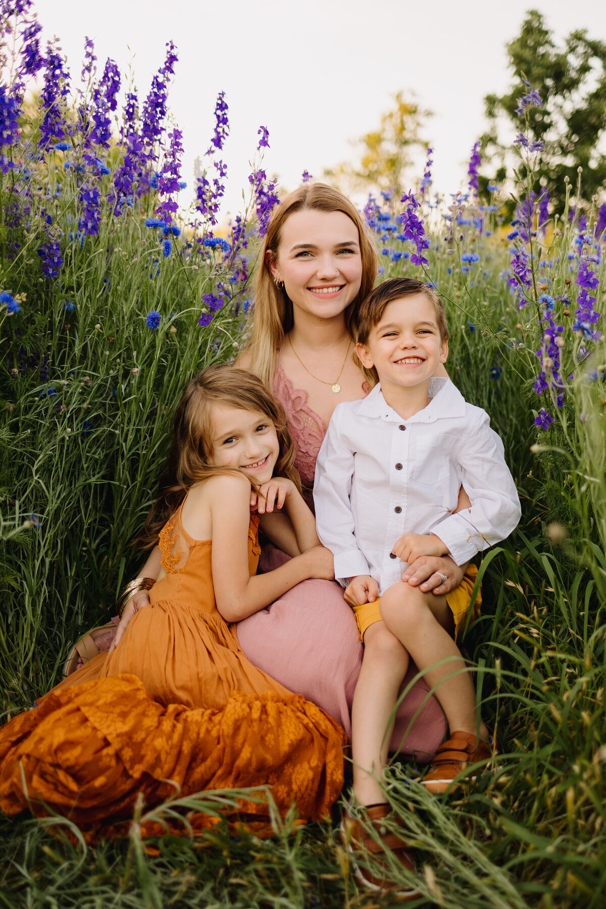 Three siblings are taking a mini photo session in spring season. They are sitting on the glass and around them are green zone  with purple flowers. The older sister is wearing a pink dress, the boy  a white shirt and the youngest girl a long orange dress.