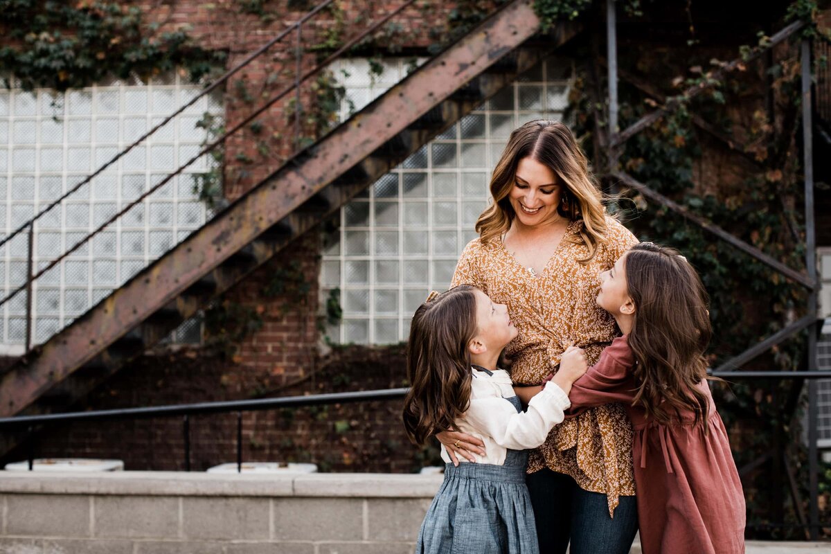 A Pittsburgh family photographer captured a woman smiling down at two young girls who are looking up at her affectionately, with stairs and a brick building in the background.