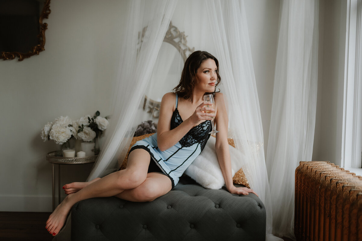 A woman in a blue slip dress poses on a couch while she looks out the window and drink champagne in the Vancouver, BC boudoir studio.