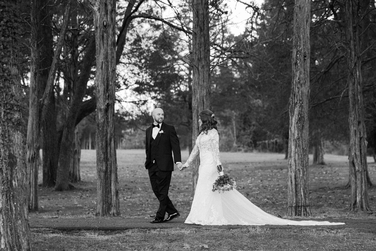 A black and white image of a bride and groom as they walk underneath tall trees
