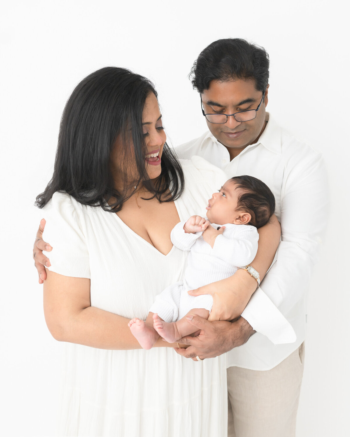 New parents wearing white look lovingly at baby in white in white studio backdrop portrait
