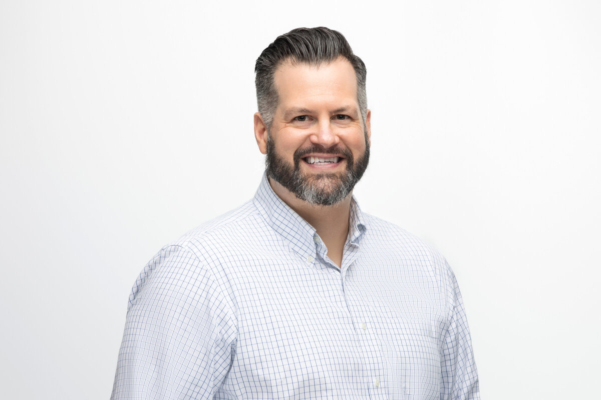 A man with dark hair and a blue and white check shirt smiles as he poses for a professional headshot portrait at Janel Lee Photography studios in Cincinnati Ohio