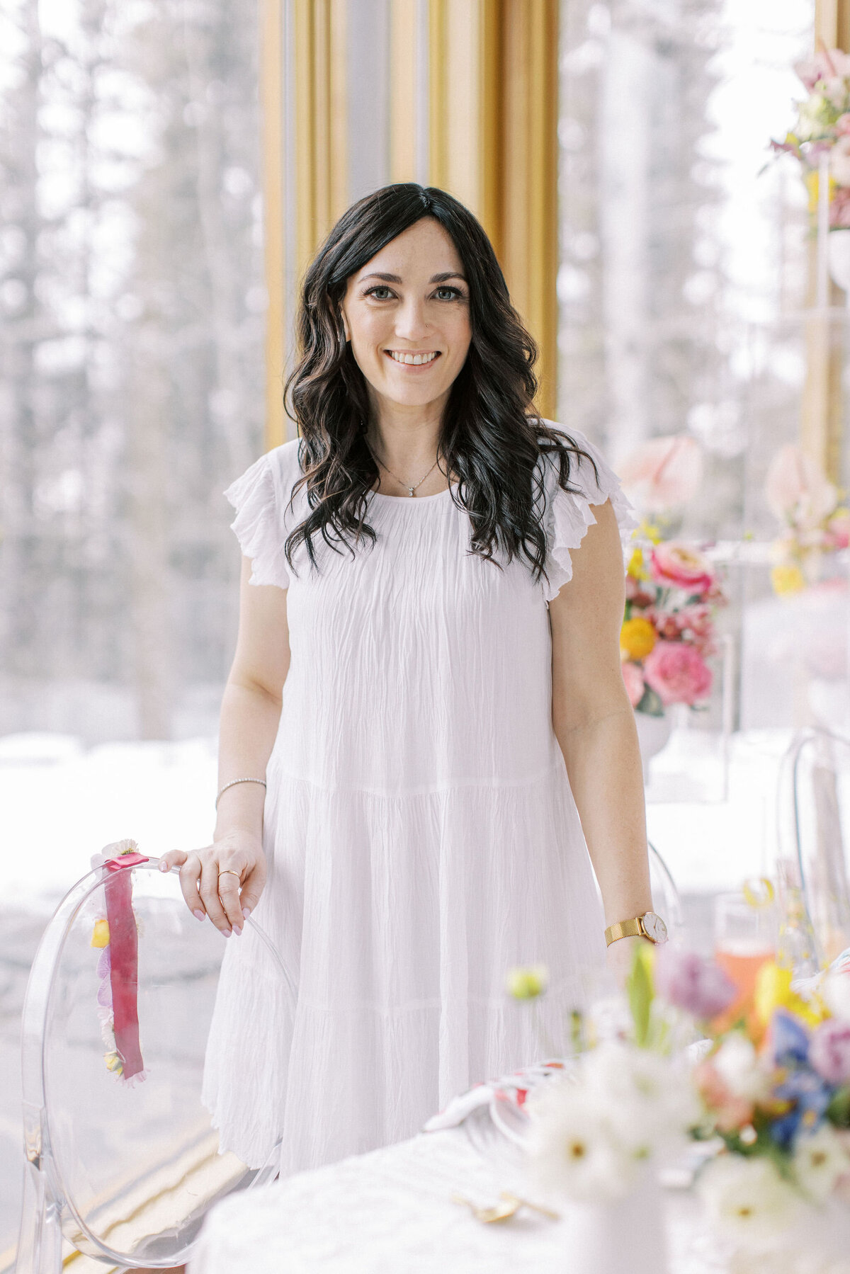 A woman wearing a white dress stands beside a table adorned with colorful flowers in a brightly lit room with large windows, capturing the essence of weddings in Banff, Alberta.