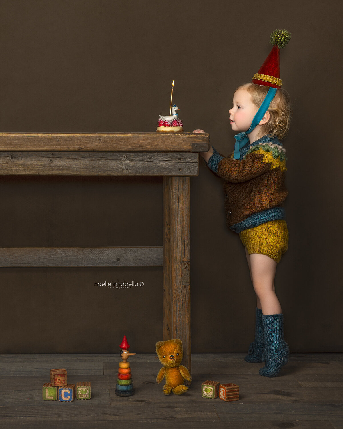 Child standing on tip toes at the edge of a table, dressed in earth tone knit clothing and a velvet party hat, blowing out a candle on a mini cake with a ceramic duck candle holder. 