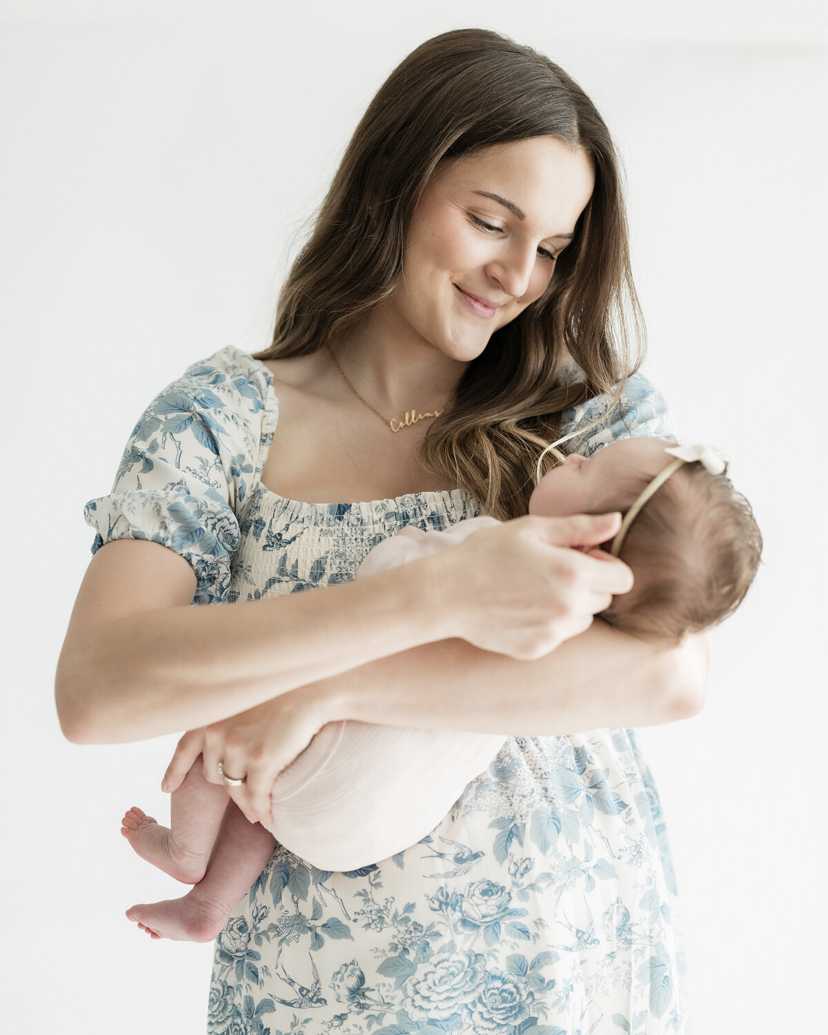 Mother in a floral dress smiling as she holds her newborn baby against a clean white backdrop during a Kansas City newborn photo session.