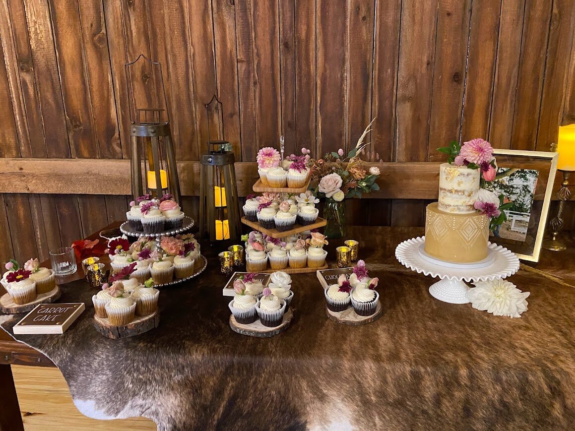 On a table with a fur style table cloth, sits an array of cupcakes with fresh flowers in shades of pinks. On the right side is a two tier cake with a tan bottom tier with stenciling, and a naked style top tier with gold leaf. There's clusters of fresh flowers in pinks. 