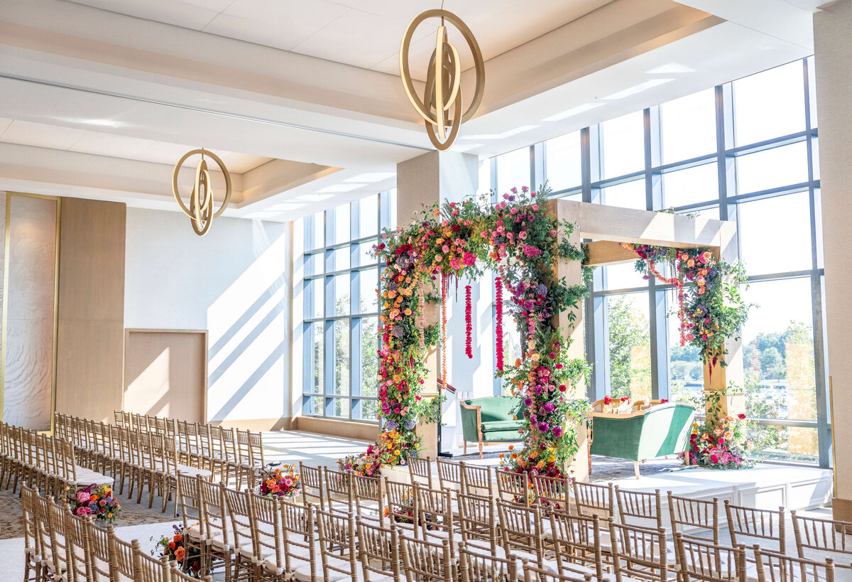 A sunlit wedding venue with rows of wooden chairs facing a floral archway. The arch is adorned with vibrant flowers in pink, red, and green. Large windows offer a view of trees outside. Elegant chandeliers hang from the ceiling.