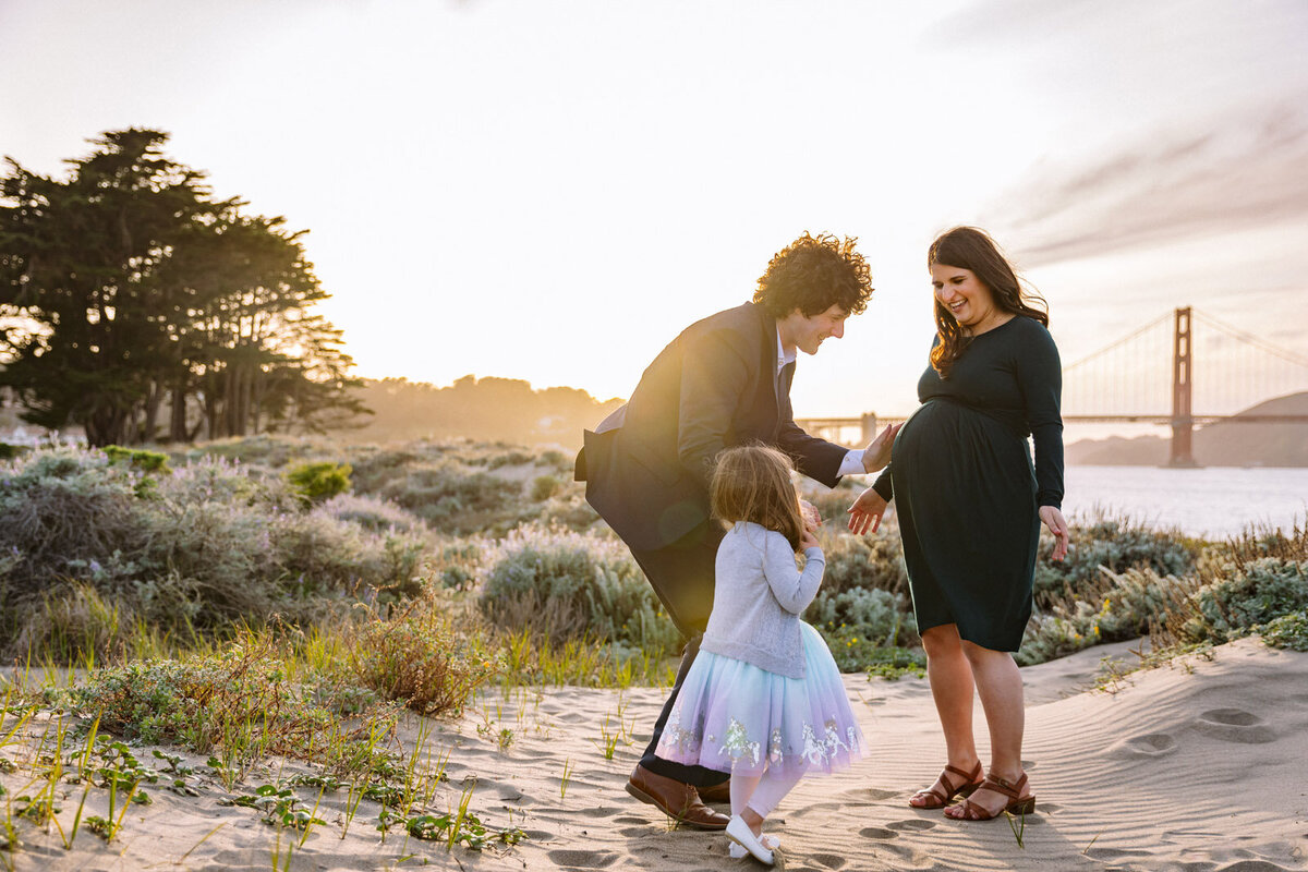 pregnant mom smiling at daughter as she and her dad go to reach for mom's baby bump at san francisco beach in front of golden gate bridge