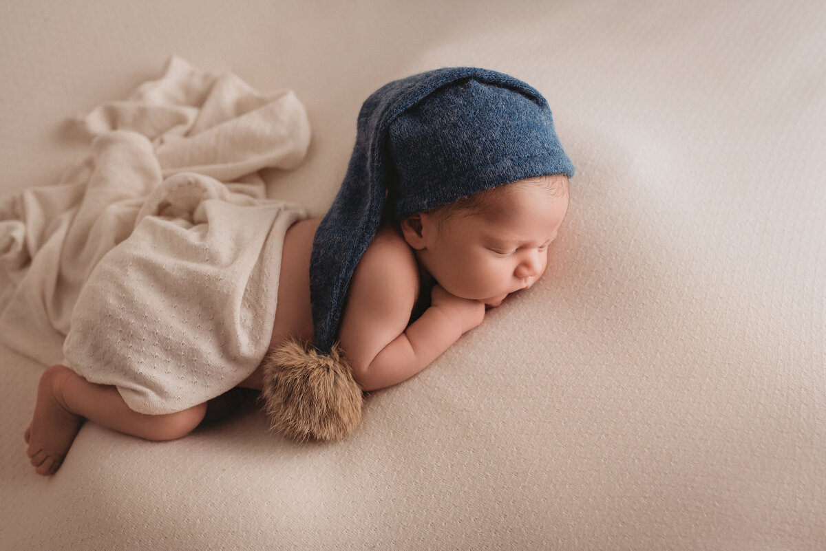 newborn baby photo session at atlanta ga newborn portait studio with one week old baby boy laying on creme fabric asleep with chin on hand wearing blue hat and cream fabric covering diaper