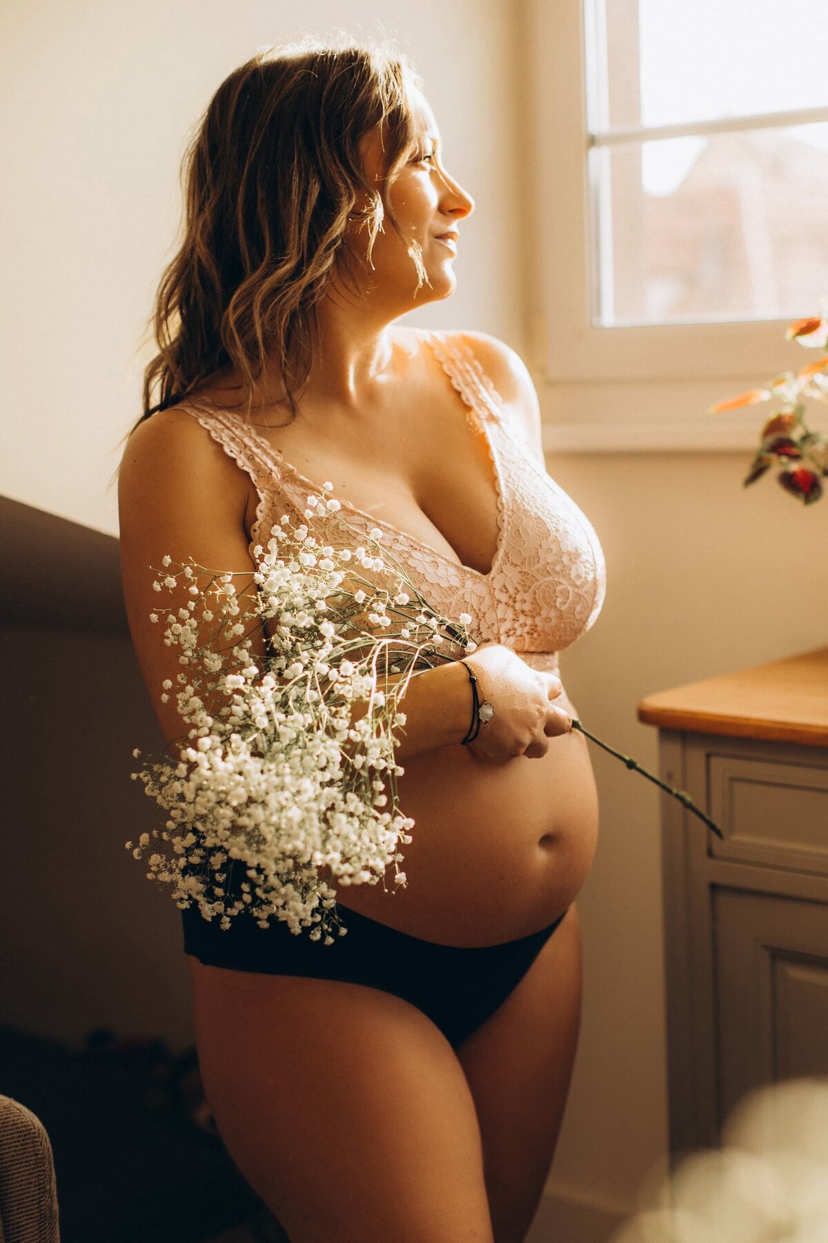 photo grossesse femme en sous-vêtement regardant par la fenêtre en tenant un bouquet de gypsophiles 