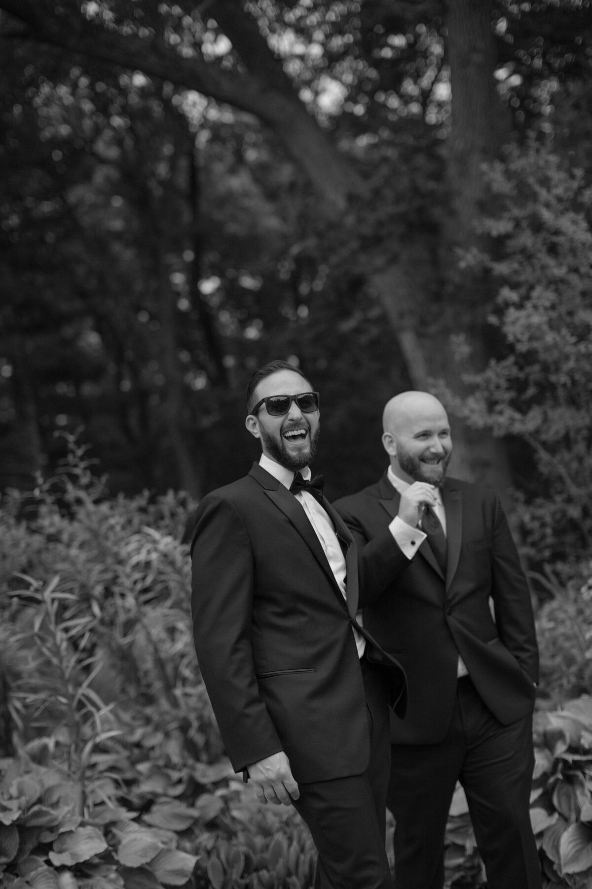 Groom laughs towards camera in black tuxedo during wedding.